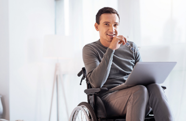 Man in a wheelchair working on a laptop with glasses in his hand smiling