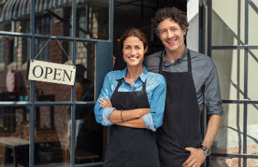 A lady and man stood outside their shop with an open sign smiling