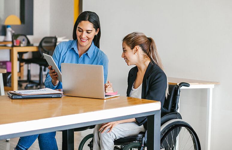 Lady in a wheelchair at a table with a woman looking at a tablet and laptop