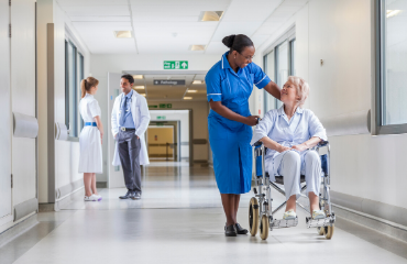 Medical lady in a blue dress pushing a lady in a wheelchair down a hospital corridor 